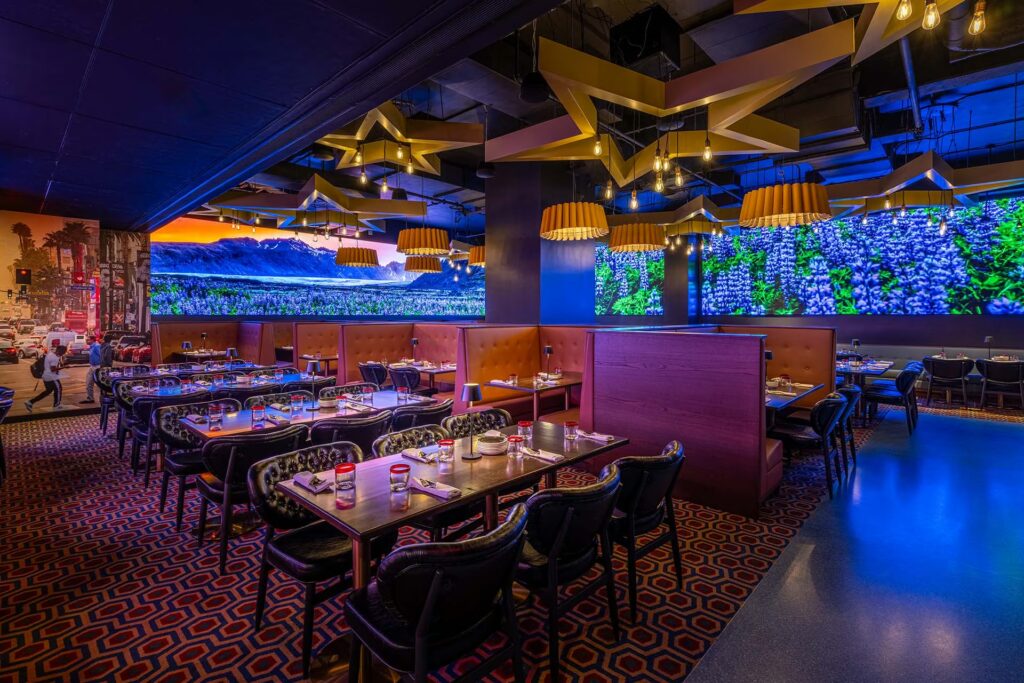 A dark, immersive restaurant interior featuring rows of tables and booths. The ceiling has star-shaped lights, and the walls are large video screens displaying a busy city street, a mountain range, and bright blue and green foliage.