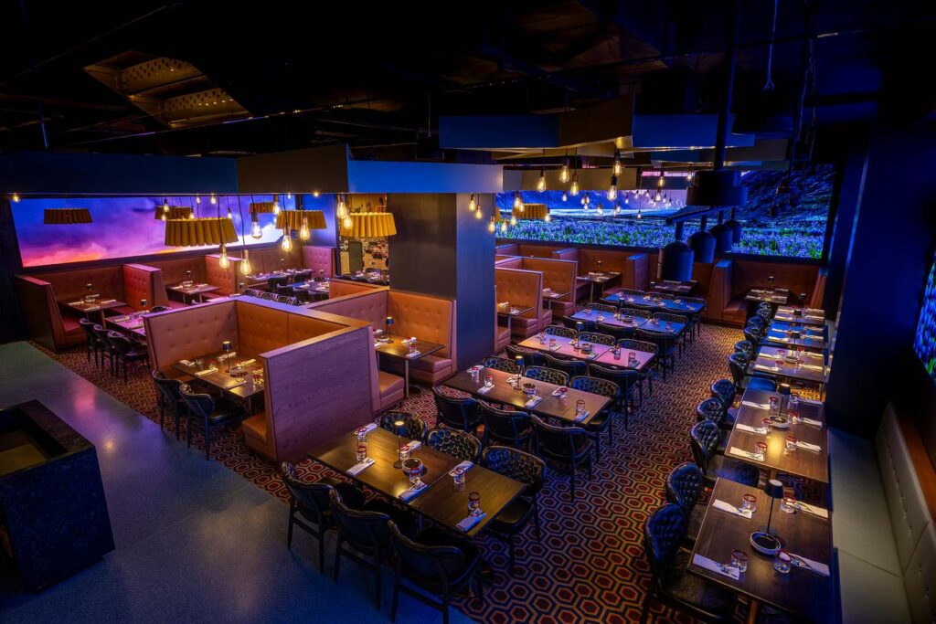 An overhead view of a dimly lit PHNYC dining room with orange booths, dark chairs, and illuminated screens displaying landscapes on the walls.