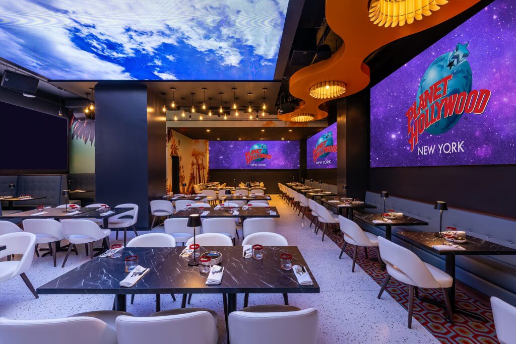A modern dining area in PHNYC with white chairs, marble tables, and a ceiling and wall screens displaying blue skies and the Planet Hollywood logo.