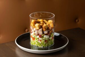A vertical chopped salad, called the Hollywood Bowl, is layered in a clear glass cylinder with lettuce, vegetables, meat, cheese, and large croutons on top.