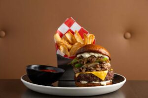A tall burger topped with cheese, sautéed onions, and peppers is served with a side of wedge fries and ketchup.