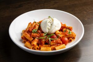 A bowl of rigatoni pasta in a red sauce with cherry tomatoes and basil, topped with a large ball of burrata cheese.