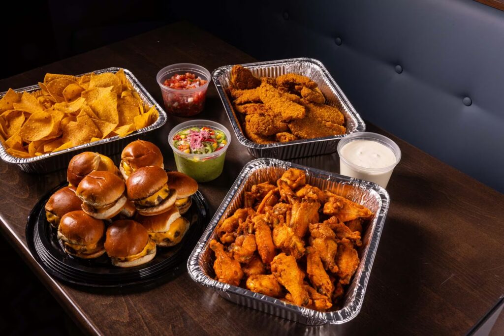 A catering display on a dark wood table features aluminum trays of buffalo wings, fried chicken tenders, tortilla chips, and a platter of mini cheeseburger sliders, with sides of salsa, guacamole, and ranch dressing.