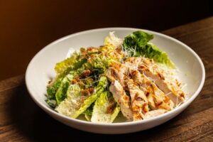 A white bowl holds a grilled chicken Caesar salad made with baby gem lettuce, cheese, and crunchy topping.