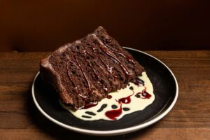 A large slice of chocolate layer cake drizzled with sauce is presented on a black plate with white and red swirled sauces.