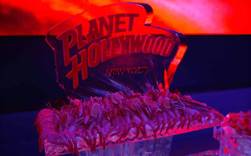 A Planet Hollywood New York ice sculpture displaying the logo and topped with a large shrimp cocktail is illuminated by red lighting.
