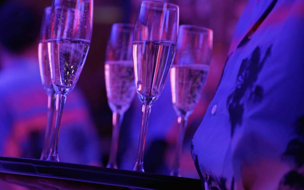 A tray of champagne flutes is carried by a server at a PHNYC event, bathed in dramatic purple and pink ambient lighting.