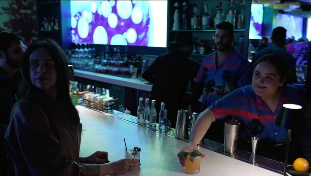 A bartender hands a cocktail to a patron over a white bar in Planet Hollywood NYC, with a purple screen displaying abstract bubbles in the background.
